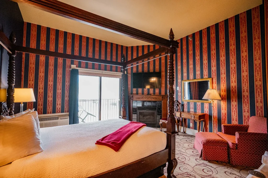 A hotel room features a four-poster bed, striped wallpaper, a fireplace with a TV, and a red armchair with an ottoman.