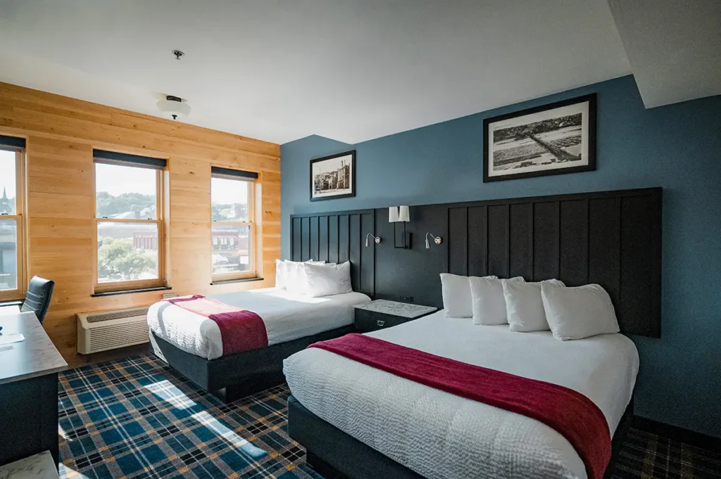 A hotel room with two queen beds, a blue plaid carpet, wood-paneled walls, and a large black headboard.