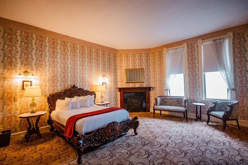 A bedroom with ornate floral wallpaper, a large wooden bed, a fireplace, two windows, and patterned carpet.