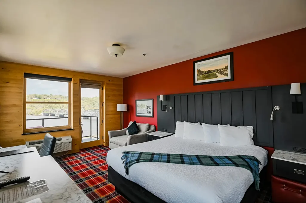 A hotel room features a large bed with a plaid runner, red walls, wooden paneled walls, and a view of a lake.