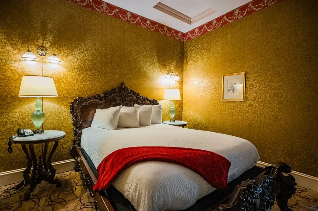 An ornate bedroom features golden damask wallpaper, a dark wood carved headboard, and a bed with a red throw blanket.