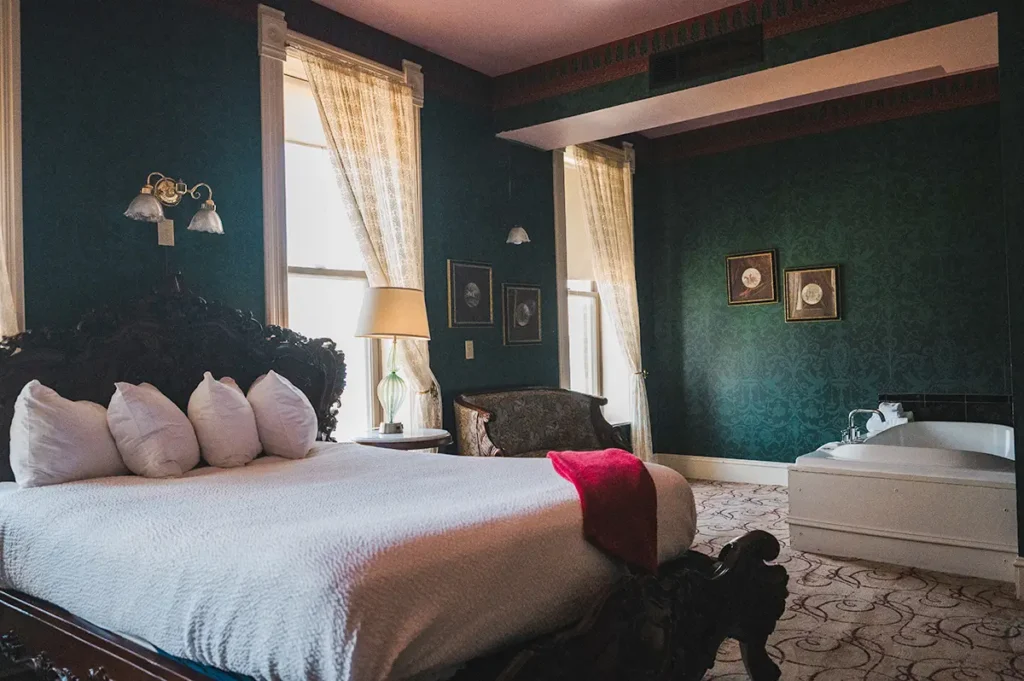 A bedroom with dark green walls, a bed with white bedding, and a white soaking tub in the corner.