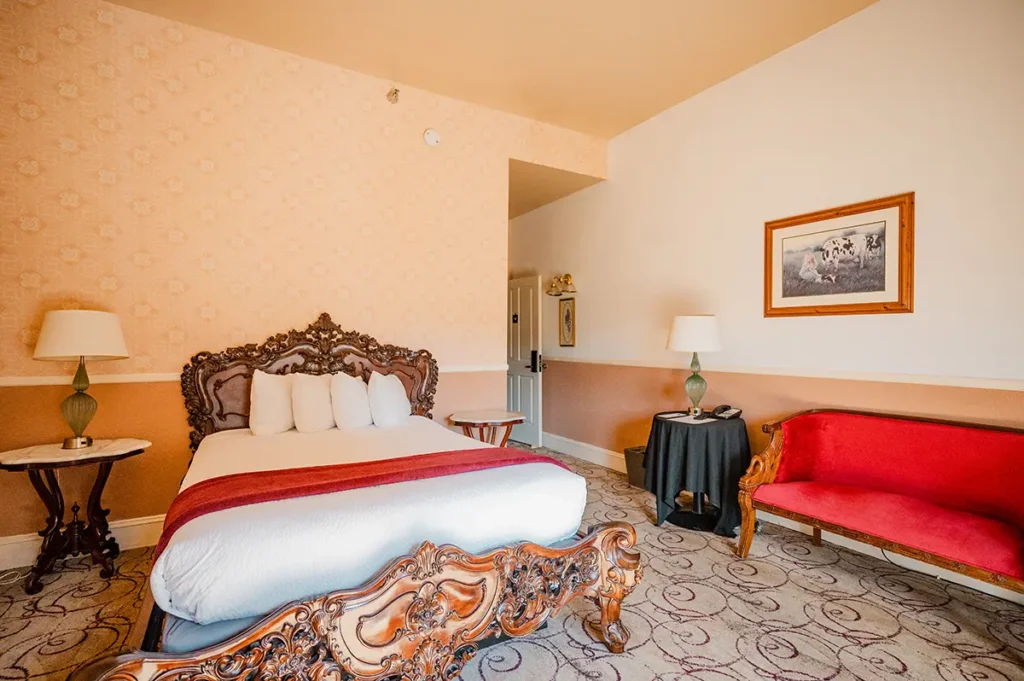 A hotel room features a large, ornate wooden bed, a red sofa, matching lamps, and patterned carpet.