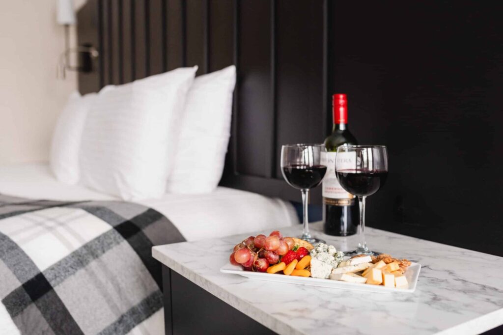 A wine bottle, two glasses of red wine, and a platter of fruit and cheese sit on a bedside table near a made bed.